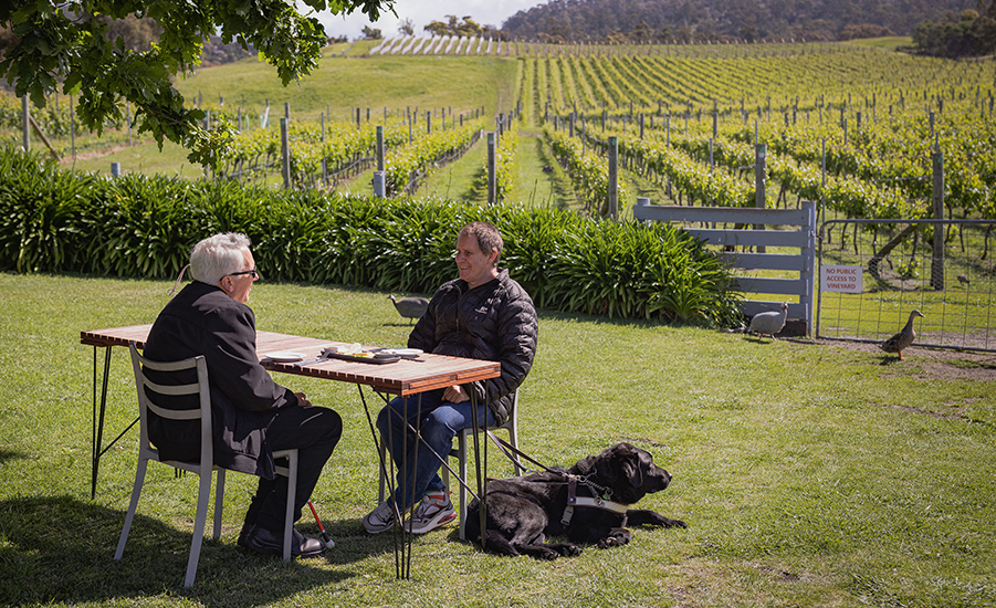 Two men are sitting at a table on the grass next to a vineyard. There is a black dog wearing an assistance harness lying on the ground next to one of the men.